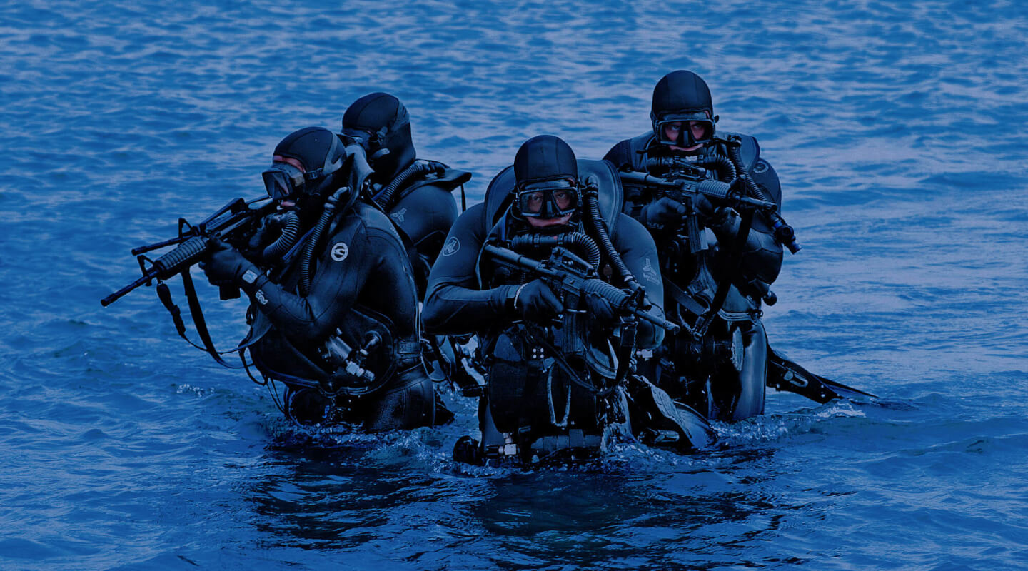 A team of four Navy SEALS, armed with rifles, emerge from or stand in the water, ready for action.