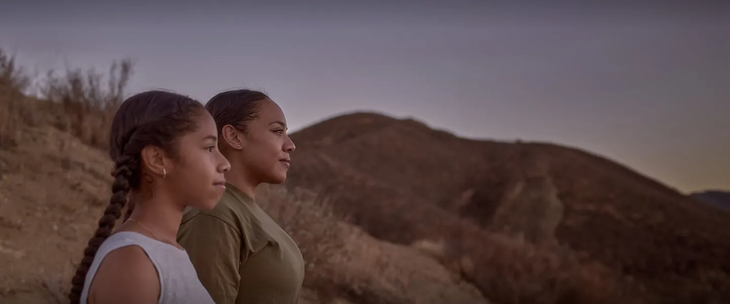 A mother and daughter stand together on a hill, silhouetted against a vibrant sunset.